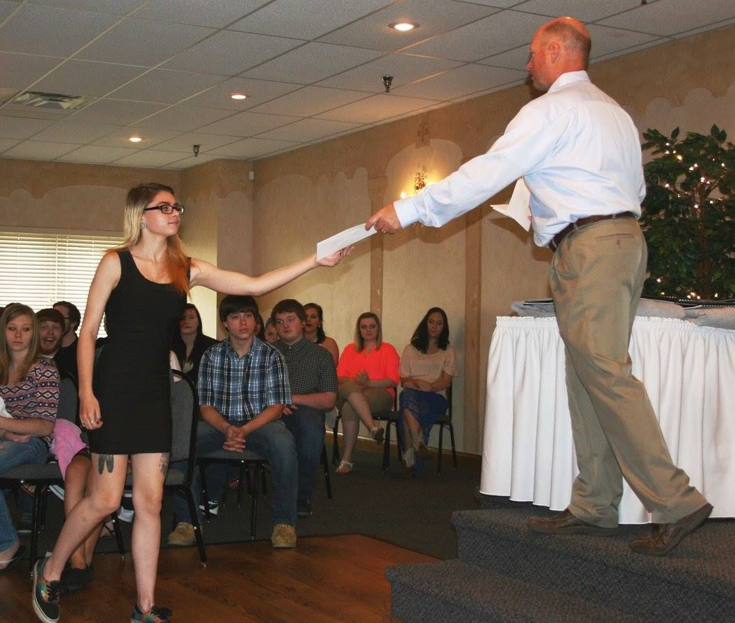 Woman in black dress receives paper from man on stage in front of seated audience.