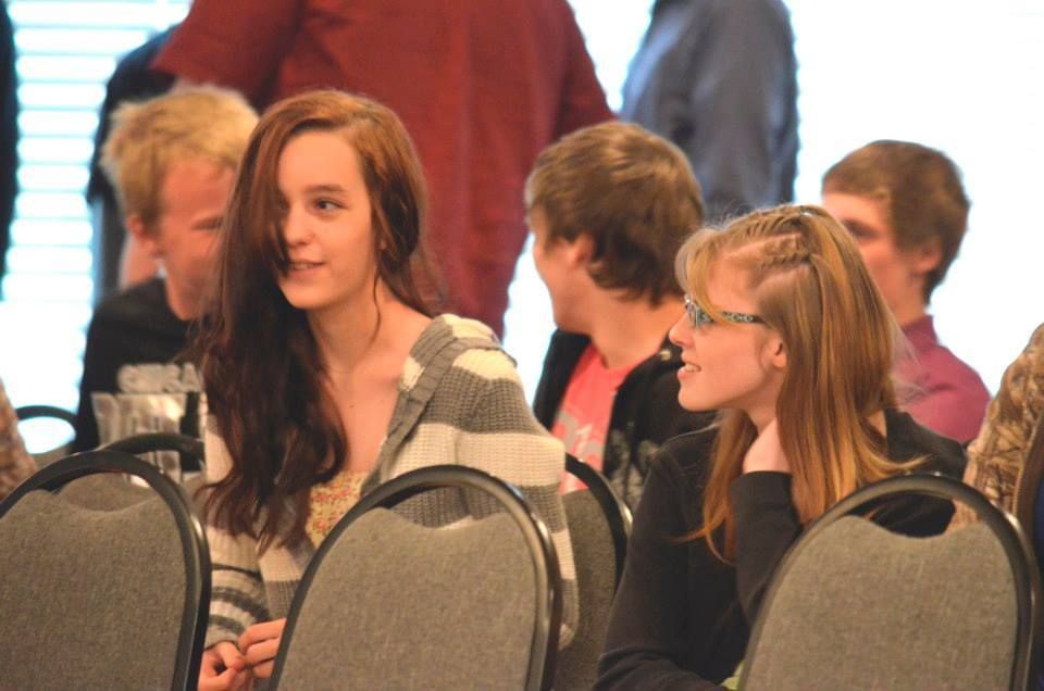 Two young people at a gathering; one in striped hoodie, the other with glasses, sitting at chairs.