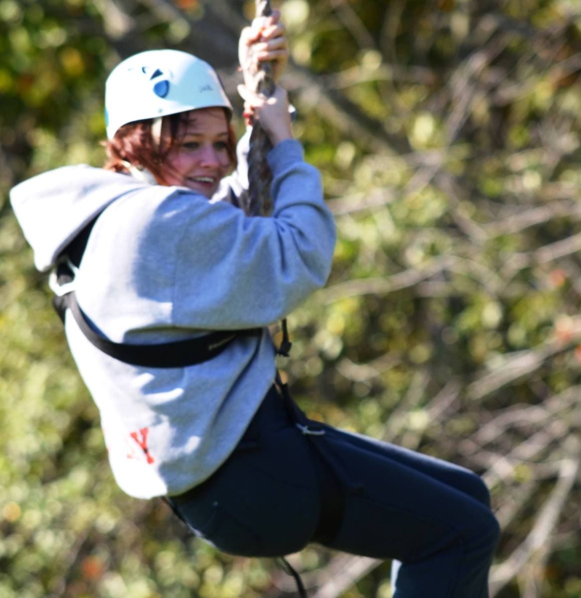 Woman on a zip line, wearing a helmet and harness, smiling outdoors.