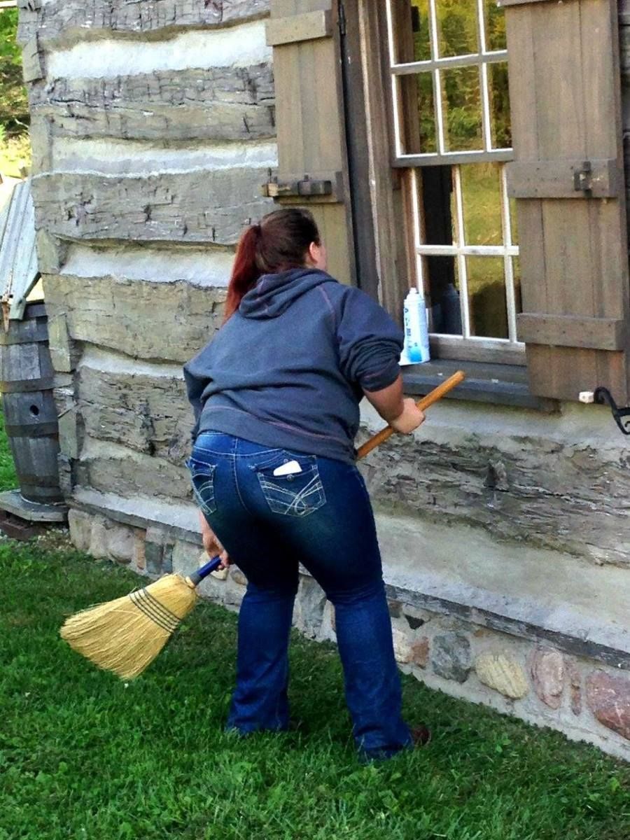 Person sweeping the side of a log cabin with a broom, near a window with shutters.