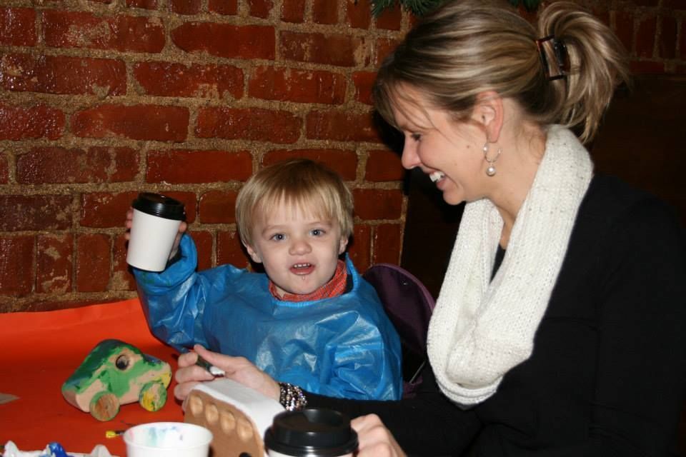 Woman smiles at a child holding a small drink. They're at a table painting.