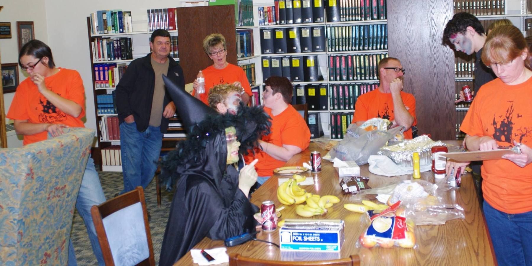 People in orange shirts around a table with a witch decoration. A library with bookshelves in the background.