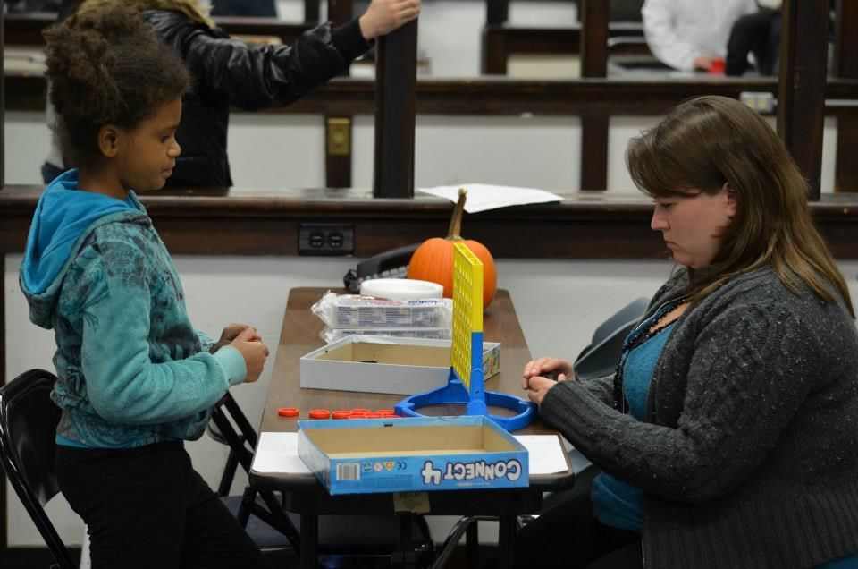 A girl and a woman playing Connect 4 at a table. A pumpkin and other items are on the table.