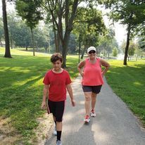 Two people walking on a paved path in a park with trees; sunny day. One wears red, the other pink.