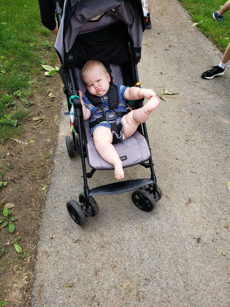 Baby sitting in a stroller, legs in the air, on a path. Person pushing stroller is partially visible.
