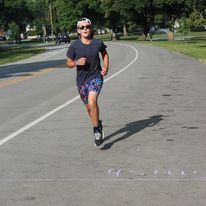 Person running on a paved road, wearing sunglasses, hat, and patterned shorts. Sunny outdoor setting.