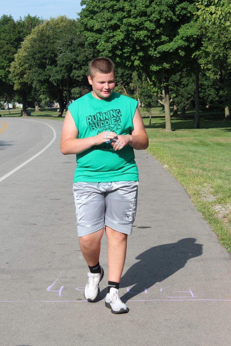 Man in green shirt and shorts walking on a path, checking watch in a park setting.