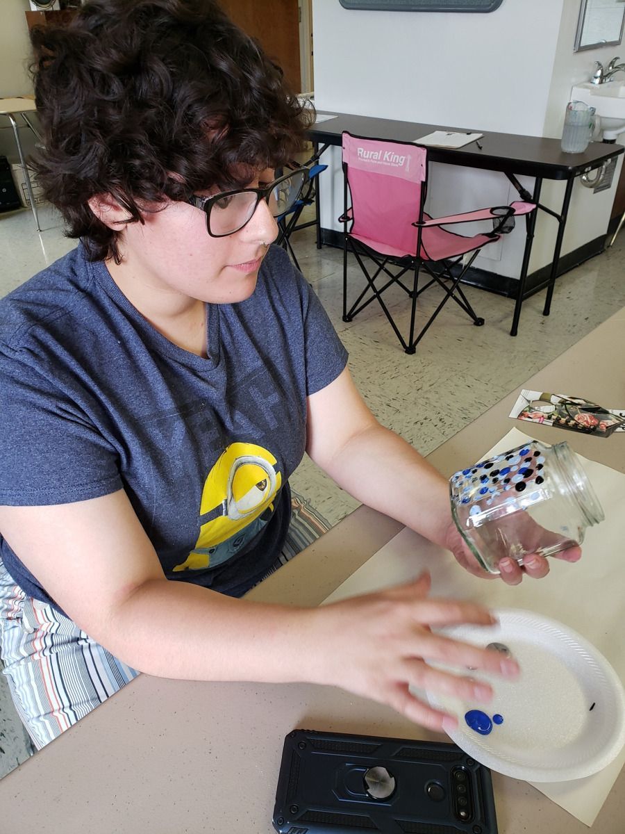 Person with glasses paints on a jar, holding it over a palette with blue paint.