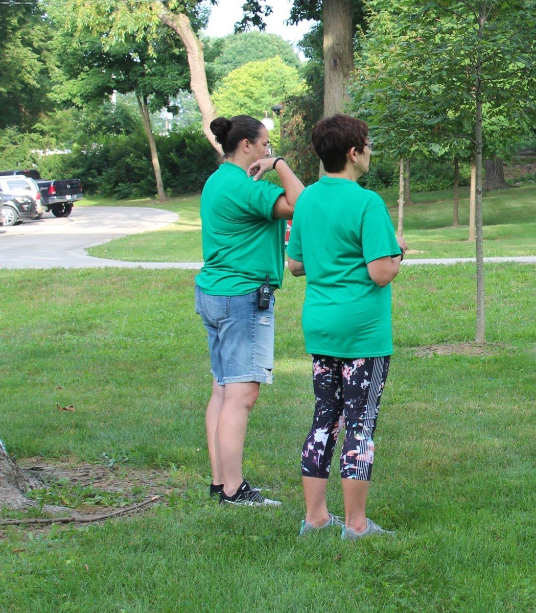 Two people in green shirts stand on grass; one adjusts the other's collar. Cars and trees are in the background.