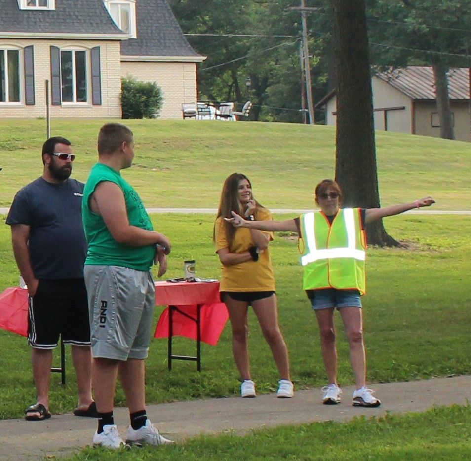 Four people standing outdoors: one directs traffic with arm extended; others watch, near a table.