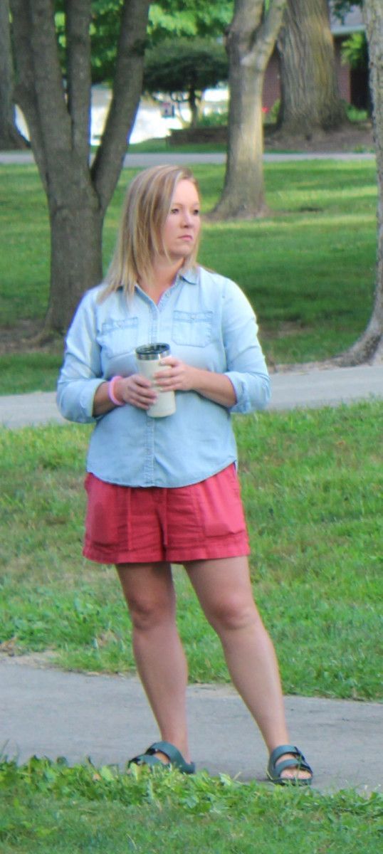 Woman in blue shirt, red shorts, holding a drink, standing outside on a path.