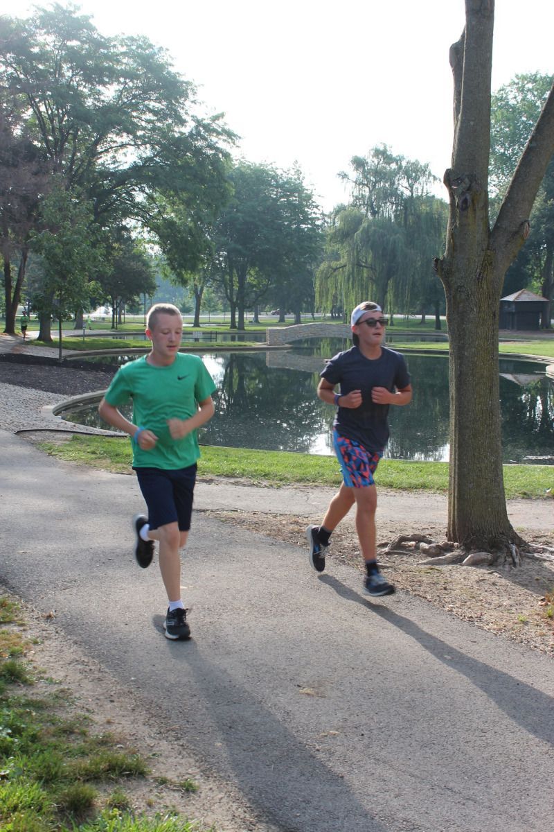 Two people running on a paved path near a pond with trees in the background.