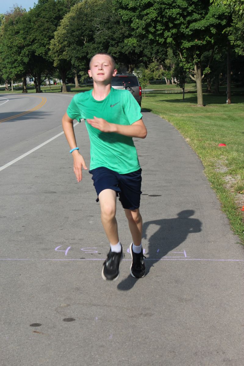Boy running on a path, wearing green shirt and blue shorts. Reaching finish line; sunny outdoor setting.