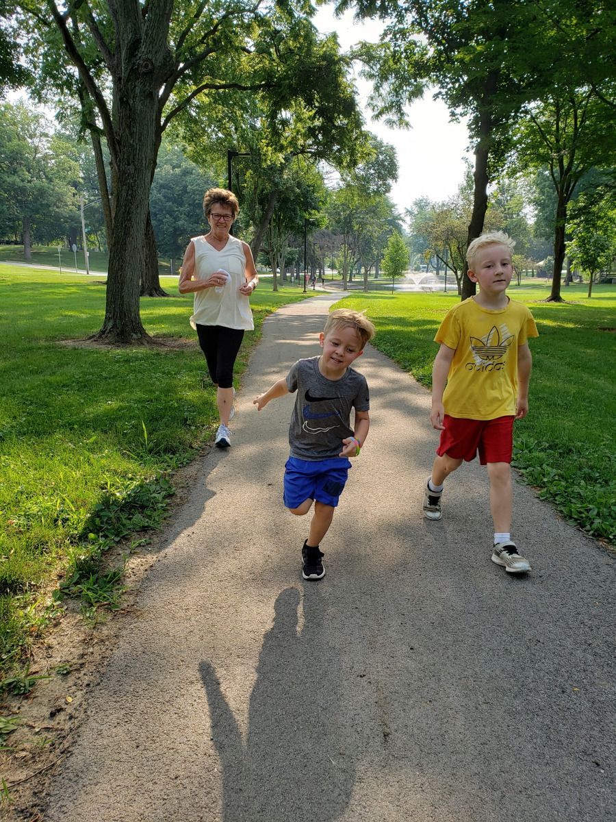 A woman and two young boys running on a paved path in a park with trees and grass.