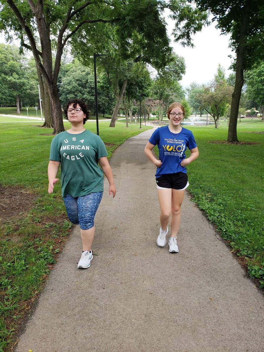Two people walking on a paved path in a park. One in blue shirt, black shorts. The other in green shirt, patterned capris.