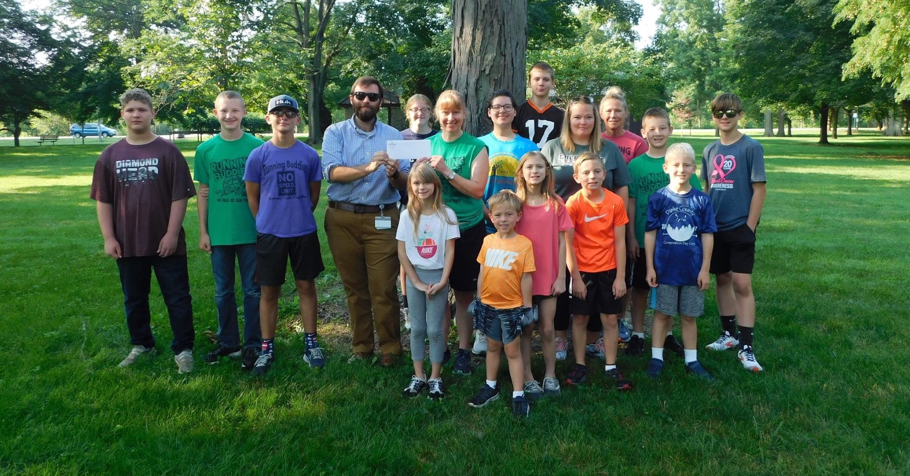 Group of people holding a check in a park, next to a large tree on a sunny day.