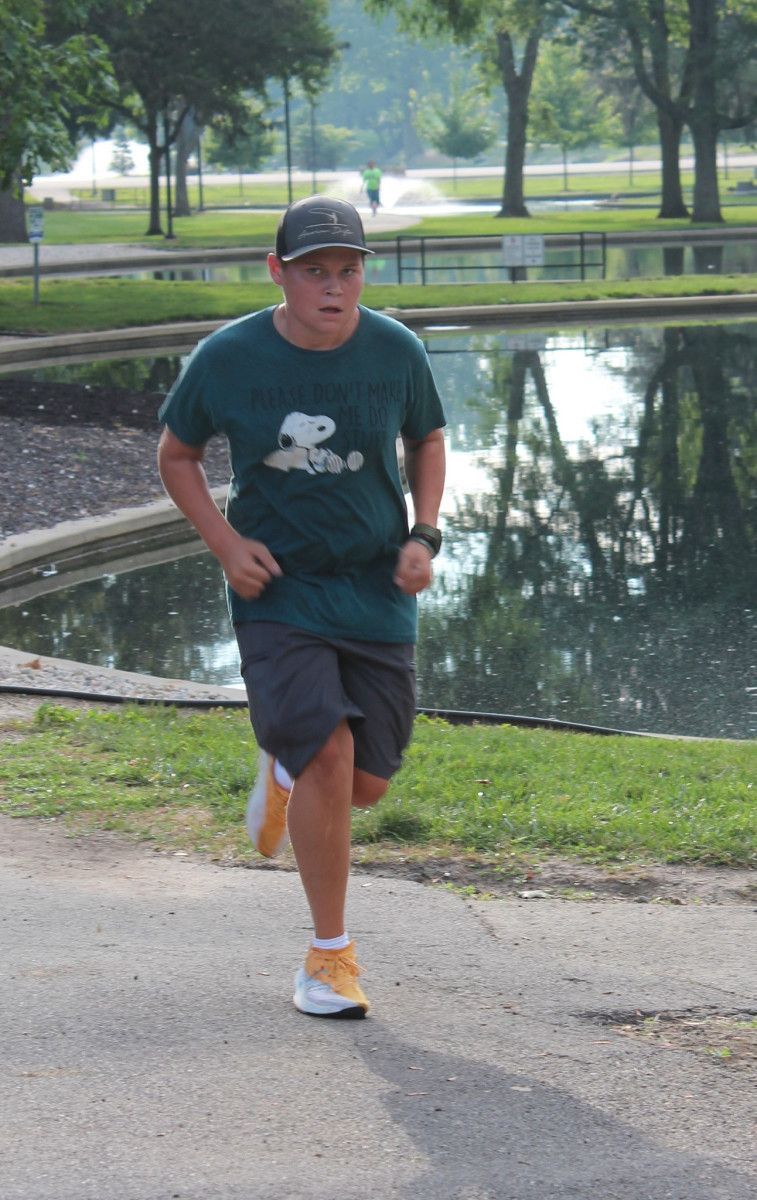 Person in a Snoopy shirt runs on a path near a pond. They wear shorts, a cap, and running shoes.