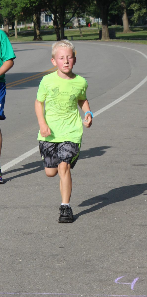 Young person running on an asphalt road; wearing a neon green shirt and patterned shorts.
