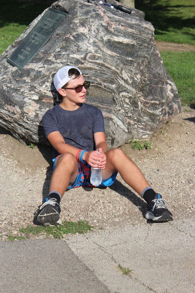 Person wearing sunglasses and a white cap, sitting and leaning against a large rock, holding water bottle.