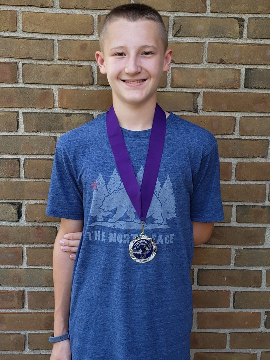 Young person smiling, wearing a blue t-shirt and purple-ribboned medal in front of a brick wall.