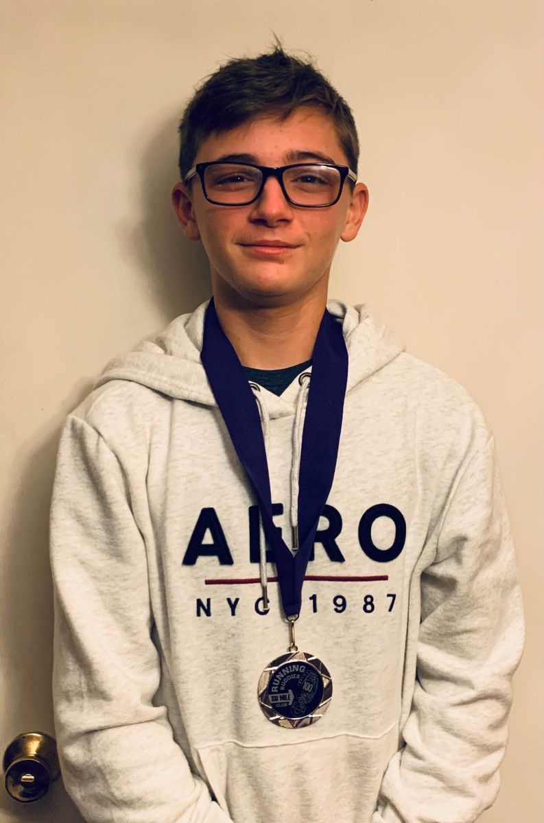 Teenager with glasses wearing a hoodie and a medal, standing in front of a neutral-colored wall.
