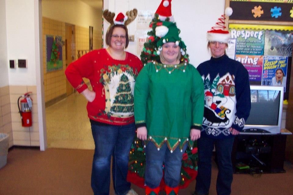 Three people in Christmas sweaters and hats posing near a decorated tree and a hallway.