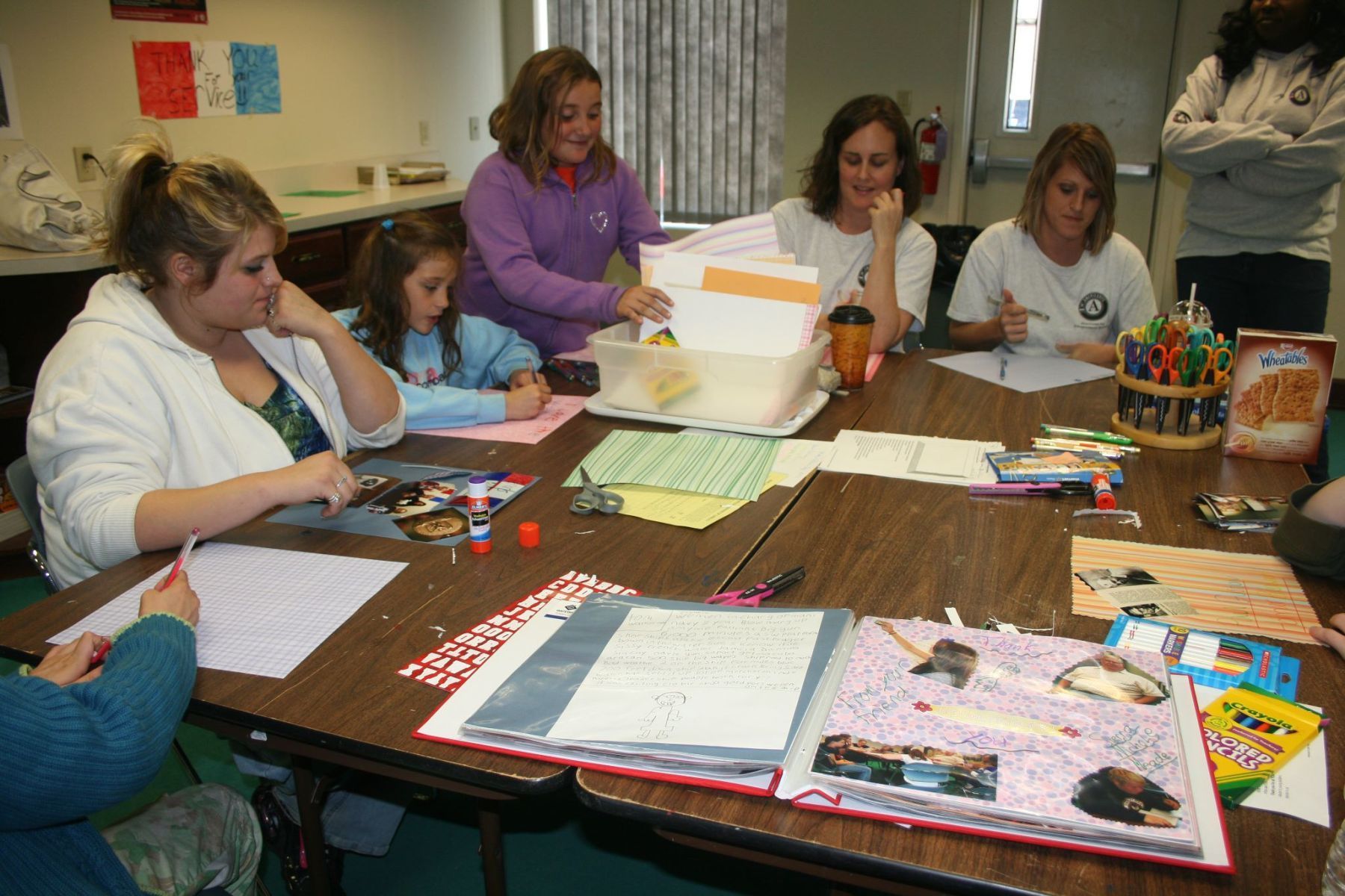 People crafting at a table: adults and a child.  Papers, glue, pens, and a scrapbook. Interior setting.