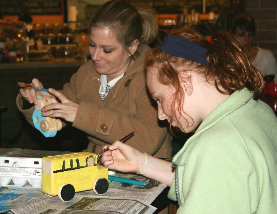 Two people painting wooden toy buses indoors; one woman wears a tan coat, the other a green shirt.