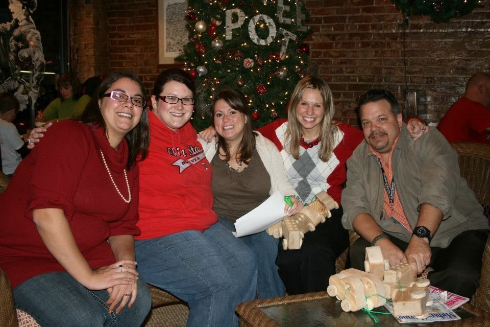 Five people, sitting and smiling, indoors near a Christmas tree. One holds a paper, another has a wooden toy.