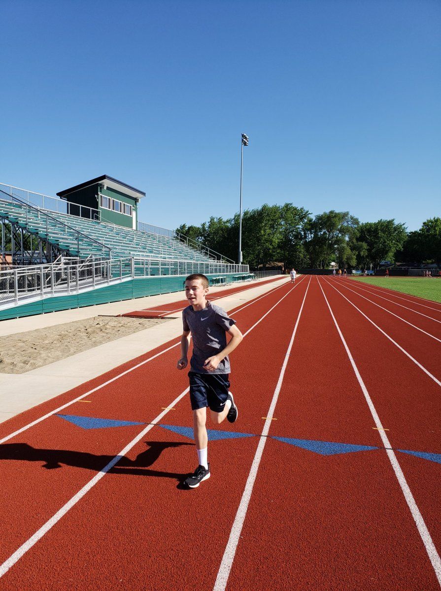 Boy running on a red track towards the camera; green bleachers and blue sky in the background.