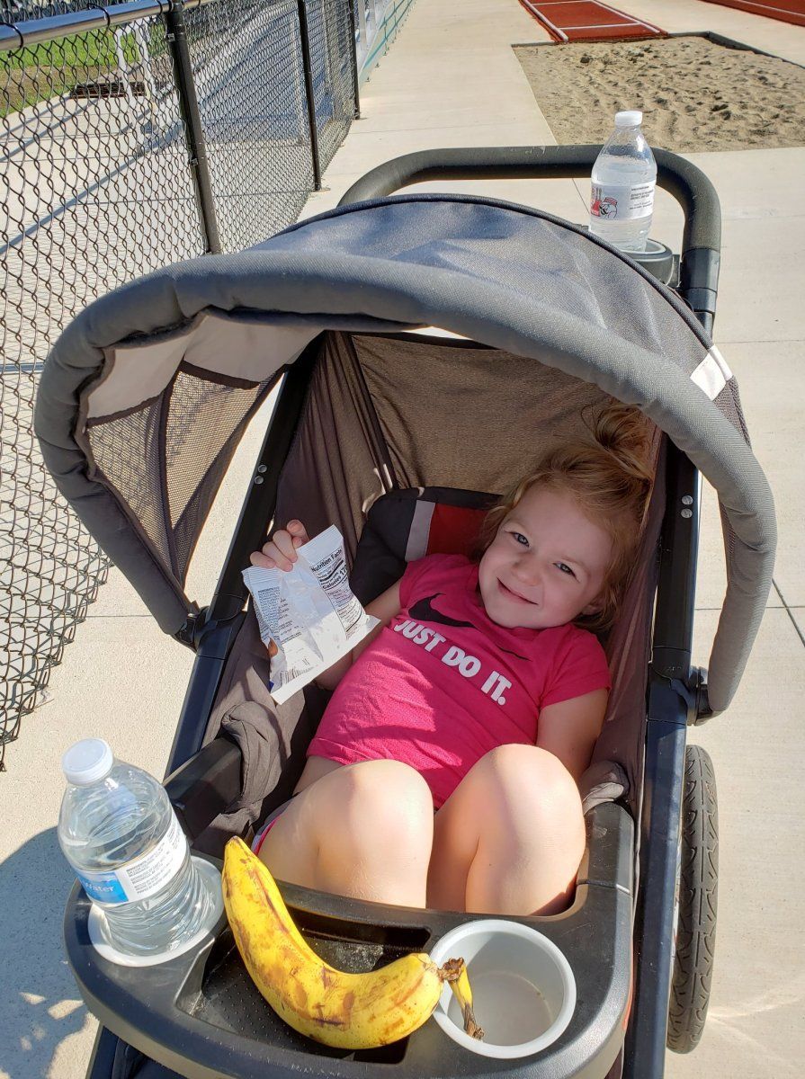 Child smiles from stroller on a sunny day; snacks and water bottles are nearby.