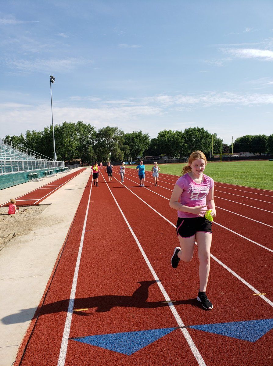 A person running on a red track toward the viewer, with other runners visible in the background on a sunny day.