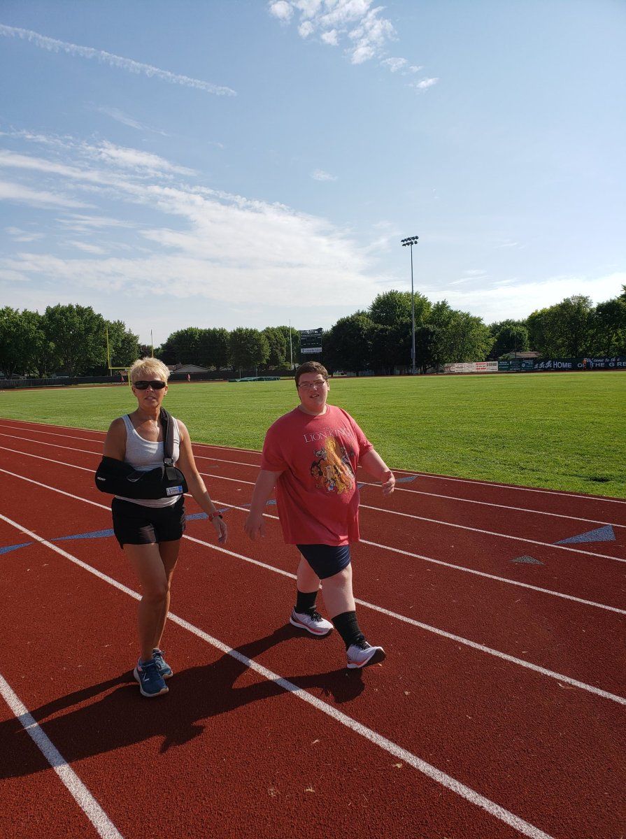 Woman with arm sling and person walk on track; blue sky, green field.