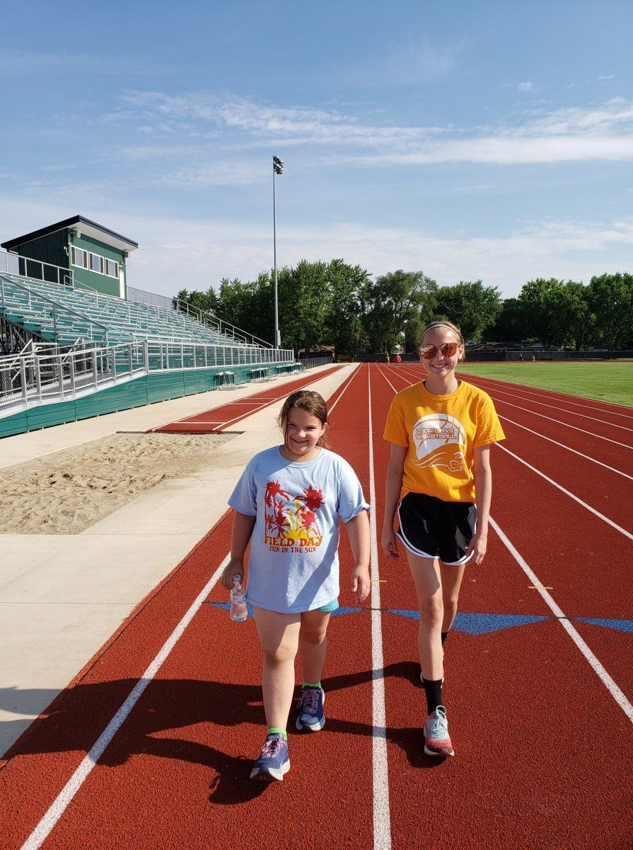 Two people walking on a red running track at a stadium on a sunny day. One wears a blue shirt, the other a yellow.