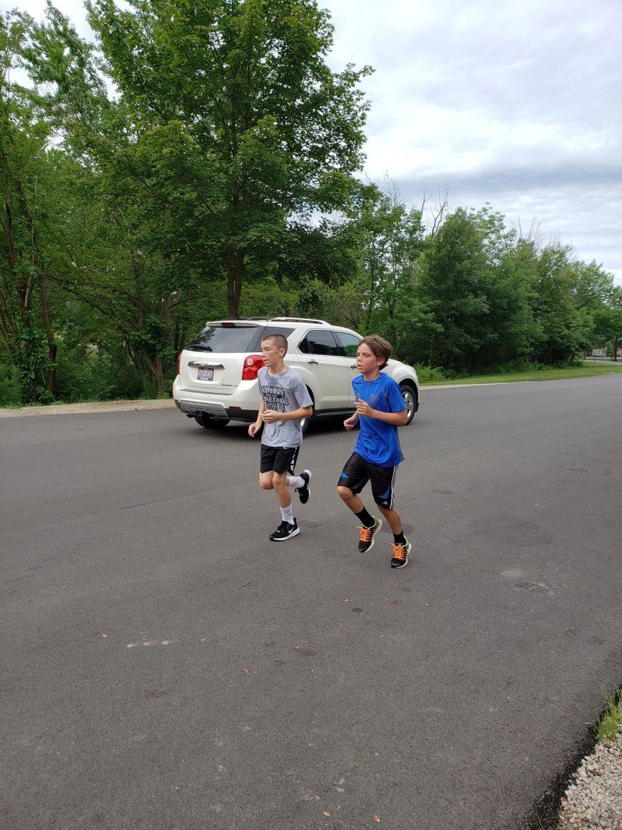 Two boys run on pavement near a parked SUV and trees. The boy on the right is in a blue shirt.