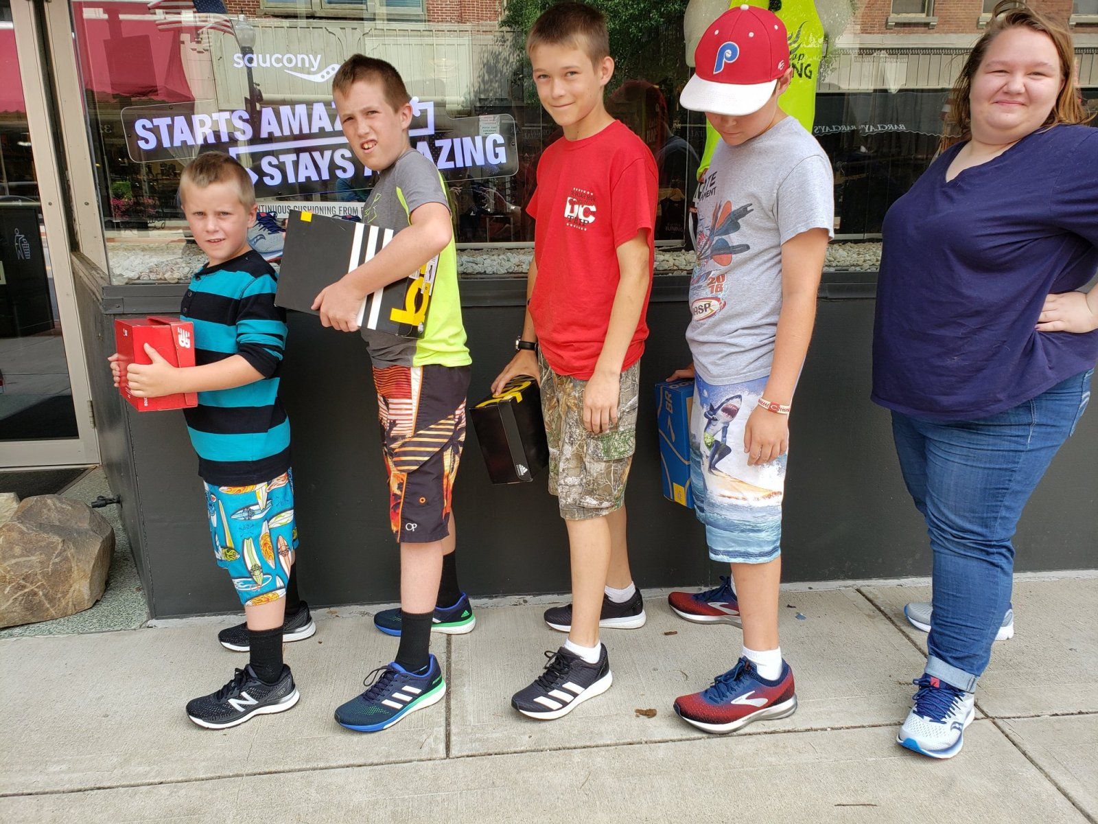 Five people standing on a sidewalk. Four boys in shorts and sneakers; a woman in jeans. Window display behind them.