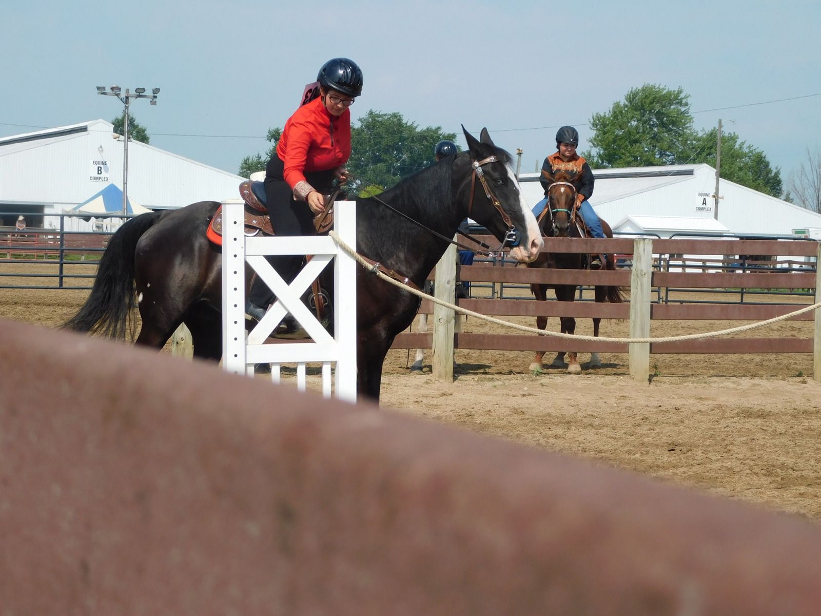 Woman on black horse near white gate, another rider behind in outdoor arena.