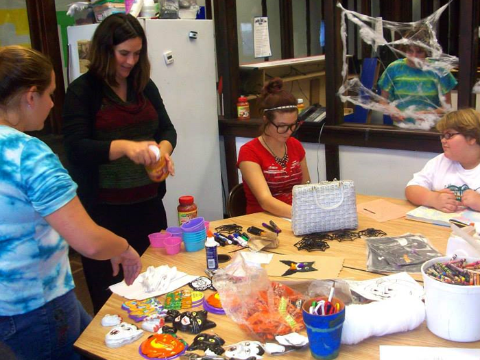 People crafting Halloween decorations at a table. Woman in black top holds a bottle. Spiders and cobwebs are visible.