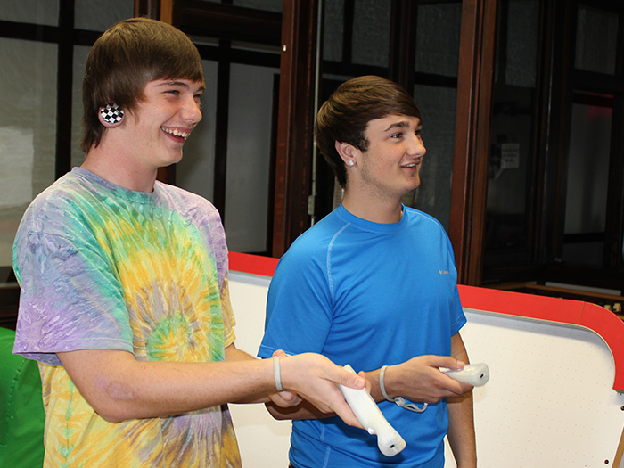 Two young men playing a video game, one laughing in a tie-dye shirt, the other smiling in a blue shirt. Indoor setting.