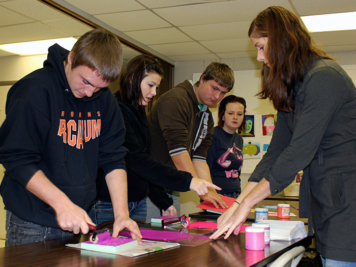 Students and teacher work on craft project at table. Pink paper and paint visible.