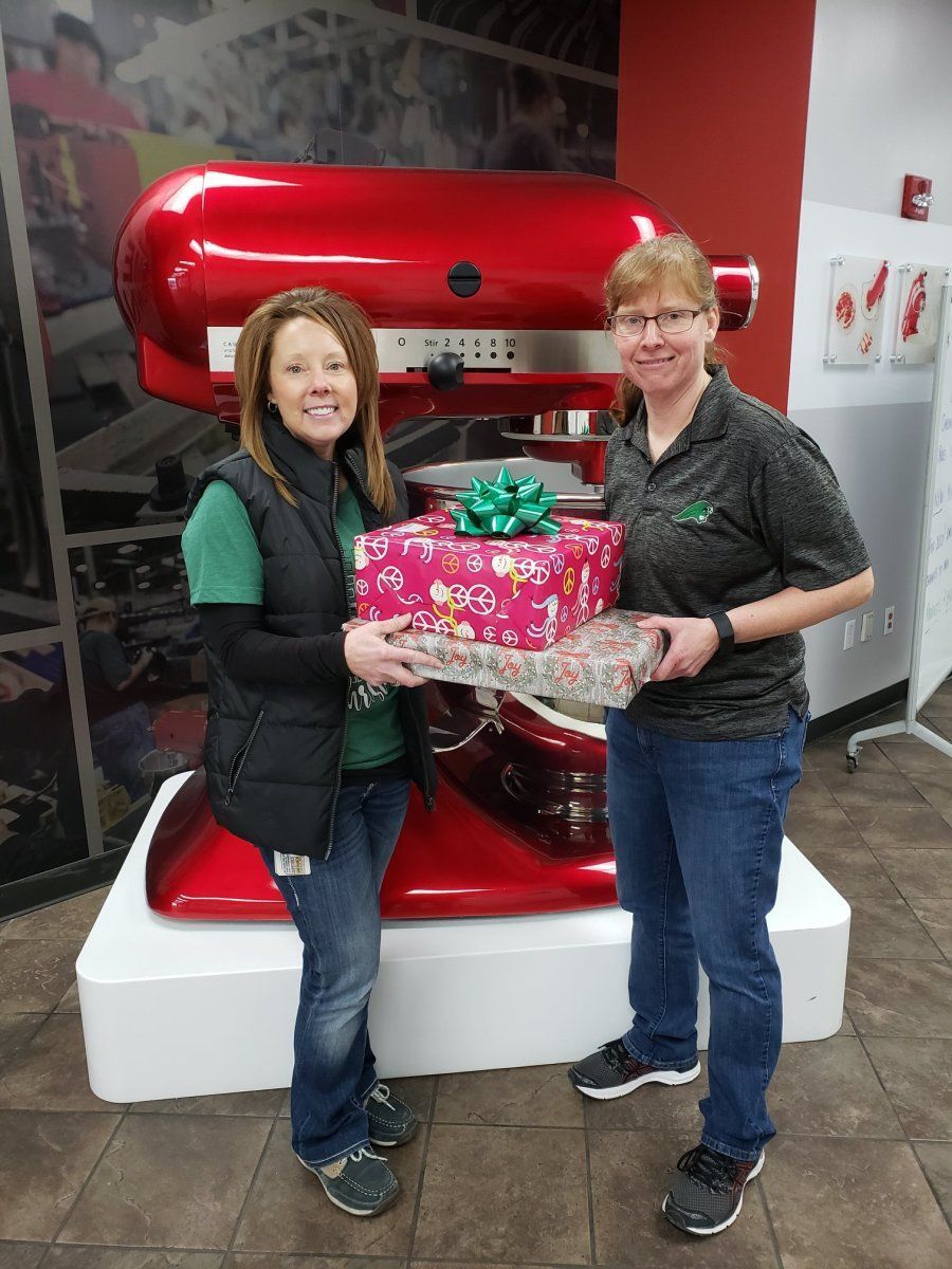 Two women hold wrapped gifts in front of a giant red KitchenAid mixer.