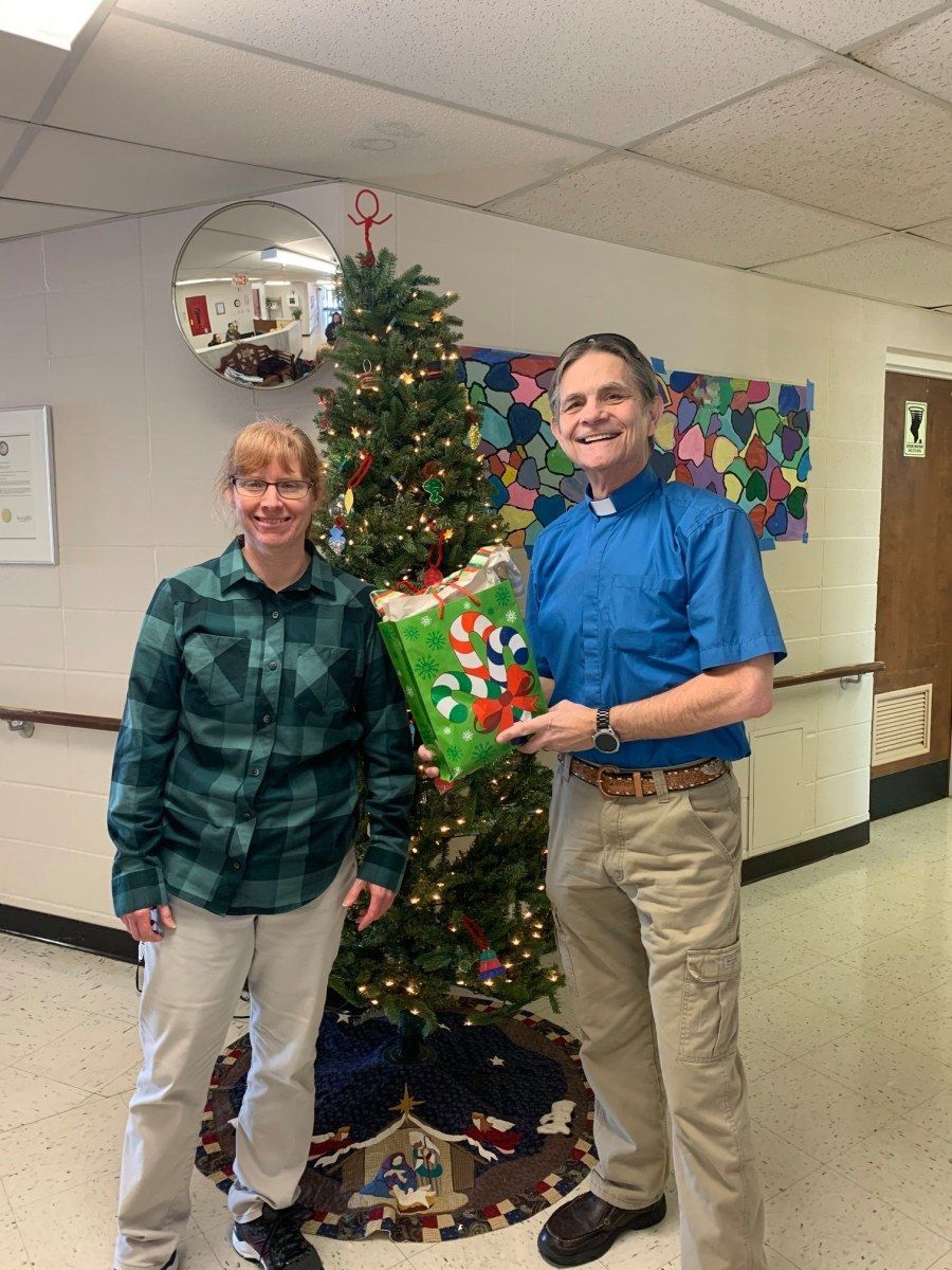 Woman and man stand by a decorated Christmas tree, holding a gift bag in a hallway.