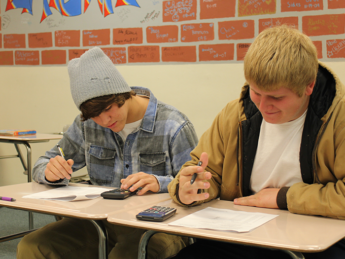 Two students work at desks in a classroom, using calculators and writing. One wears a blue beanie, the other a brown jacket.