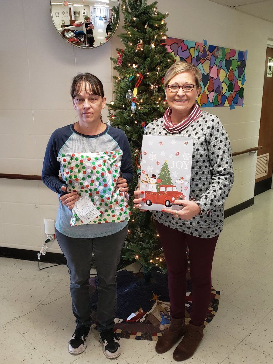 Two women holding wrapped gifts by a decorated Christmas tree in a hallway.