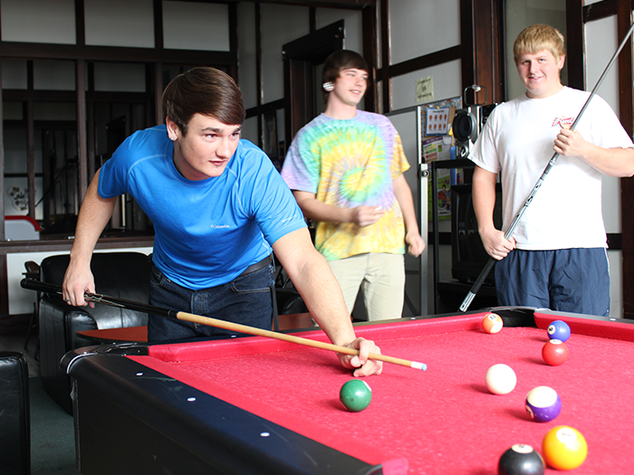 Three people playing pool in a room; one shooting, two watching. Red pool table, colorful balls.