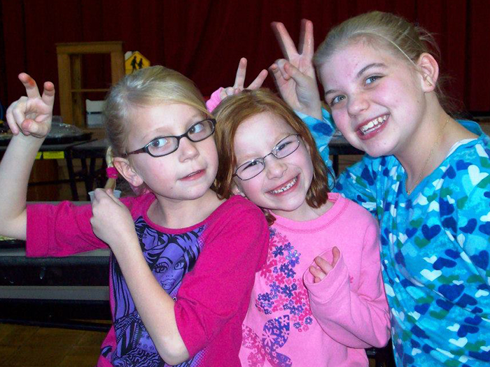 Three girls smiling and making peace signs indoors, wearing glasses and colorful shirts.