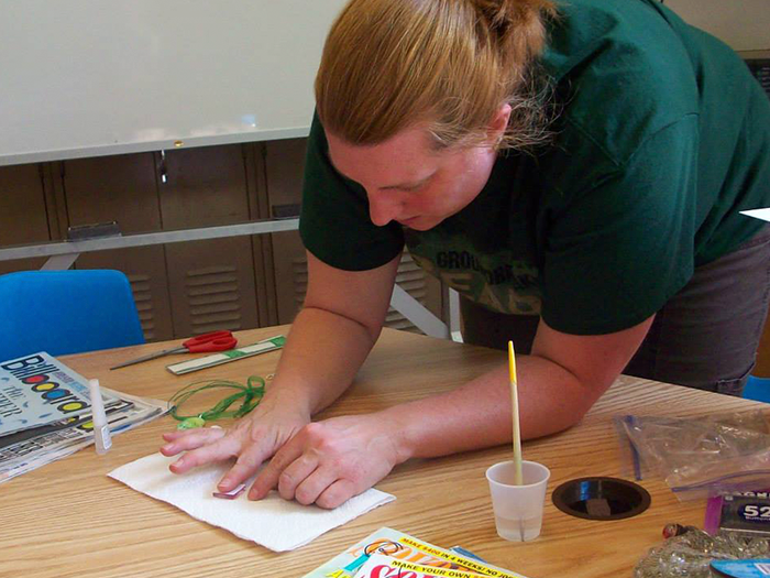 Woman in green shirt leans over a table, applying something to a paper with her finger.