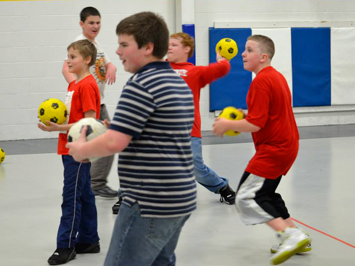 Boys in a gym play with balls, preparing to throw. Some wear red shirts.