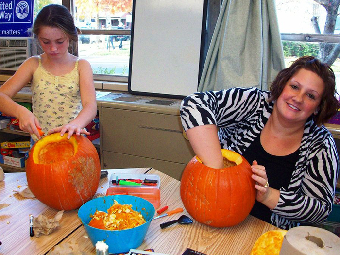 Girl and woman carving pumpkins at a table. Woman smiles, reaching inside her pumpkin; girl carves her own.