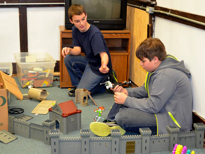 Two people playing with toy castles and figures in a room with a TV and storage.